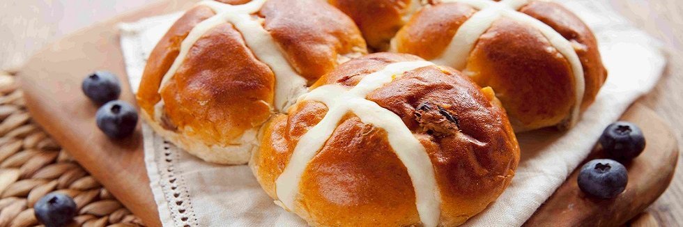Auf einem Holzbrett liegen vier hellbraune weich aussehende Brötchen. Jedes hat ein helles Kreuz aus hellem Teig eingebacken. Als Dekoration ein Deckchen und Blaubeeren.