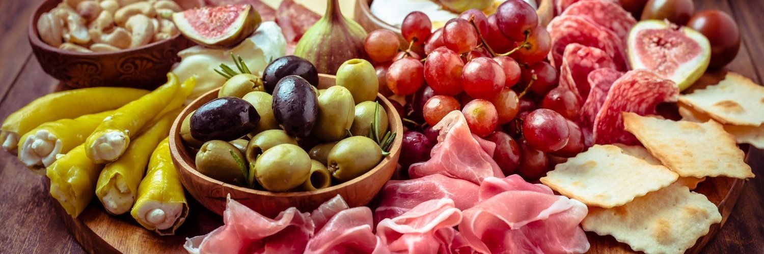 Auf einem runden Holzbrett sind Schinkenscheiben, Käse, Oliven, Trauben, Cracker, Feigen, gefüllte Peperoni und Nüsse angerichtet.