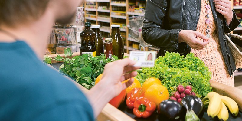 Verkäufer an der Kasse kontrolliert Mitgliedsausweis eines Genossenschaftsmitglieds. Auf dem Kassenband liegt buntes Obst und Gemüse. Dahinter eine einkaufenden Person.. Artikel "Erzeuger-Verbraucher-Genossenschaften" öffnen.