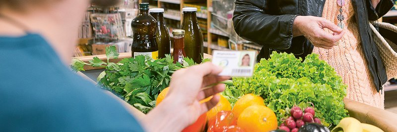 Verkäufer an der Kasse kontrolliert Mitgliedsausweis eines Genossenschaftsmitglieds. Auf dem Kassenband liegt buntes Obst und Gemüse. Dahinter eine einkaufenden Person.