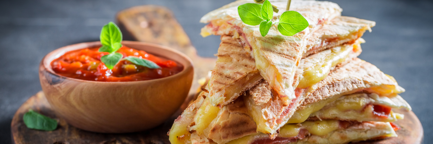 Auf einem Holzbrett liegen in Dreiecke geschnittene Quesadillas übereinander. Daneben steht ein Schüsselchen mit roter Salsa.
