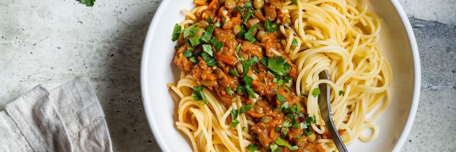 Auf einem weißen Teller ist vegetarische Linsen-Bolognese mit Pasta und Petersilie angerichtet.