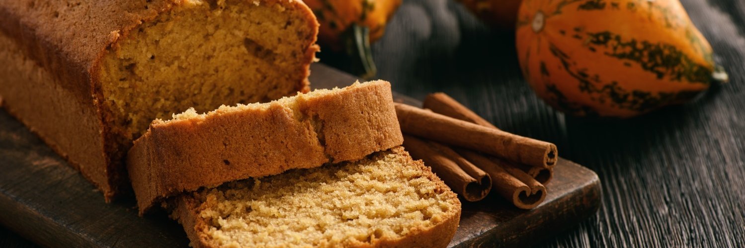 Auf einem dunklen Holztisch liegt ein kastenförmiges aufgeschnittenes Kürbisbrot, daneben Zimtstangen und Zierkürbisse.