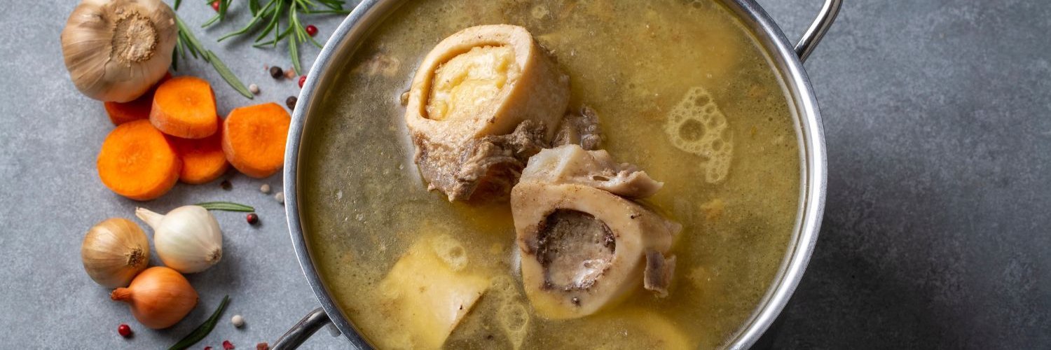 In einem Topf schwimmen Knochenstücke in einer Brühe. Neben dem Topf liegen Zwiebeln, Knoblauch, Möhren und Gewürze.
