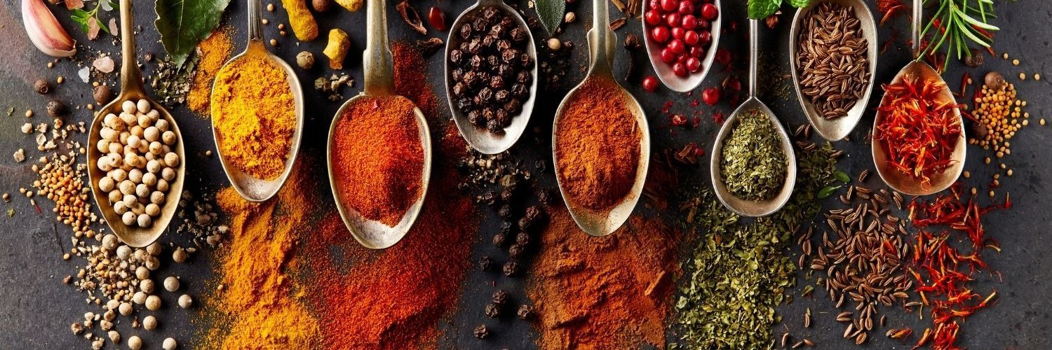 Auf einer dunklen Platte liegen neun Löffel nebeneinander mit verschiedenen Gewürzen darauf, etwa Pfefferkörner, Kurkuma, Paprika, Sternanis, Knoblauch und Rosmarin.