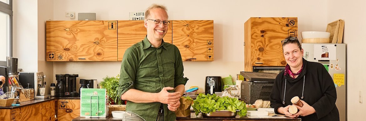 Ein Mann und eine Frau stehen in einer Kueche und laecheln in die Kamera.