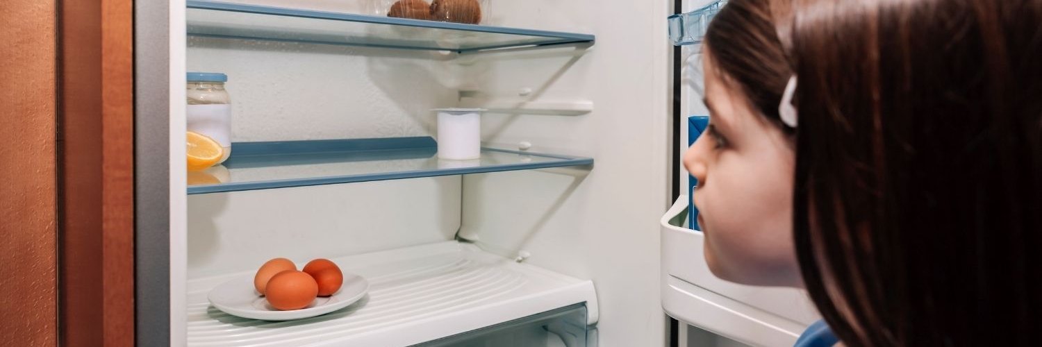 Ein dunkelhaariges Mädchen im Grundschulalter öffnet einen Kühlschrank und schaut sich dessen spärlichen Inhalt an.