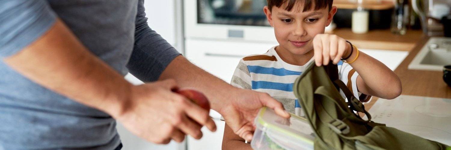 Ein Vater steckt seinem Sohn die Brotdose und einen Apfel in die Schultasche. 