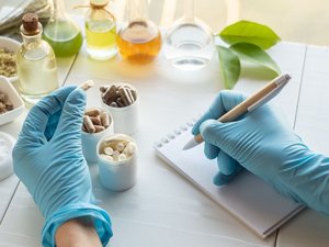Abgebildet sind zwei Hände mit Einmalhandschuhen. In der einen Hand ist eine Nahrungsergänzungsmittel-Kapsel und mit der rechten Hand werden Notizen gemacht. Im Hintergrund sind Tabletten und Gläser mit verschiedenen Flüssigkeiten zu sehen.