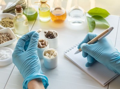 Abgebildet sind zwei Hände mit Einmalhandschuhen. In der einen Hand ist eine Nahrungsergänzungsmittel-Kapsel und mit der rechten Hand werden Notizen gemacht. Im Hintergrund sind Tabletten und Gläser mit verschiedenen Flüssigkeiten zu sehen.