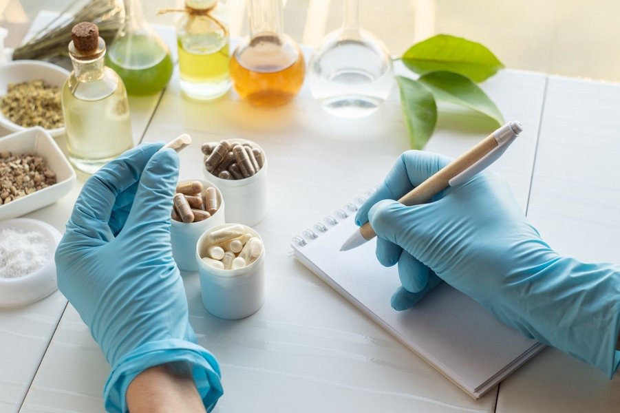 Abgebildet sind zwei Hände mit Einmalhandschuhen. In der einen Hand ist eine Nahrungsergänzungsmittel-Kapsel und mit der rechten Hand werden Notizen gemacht. Im Hintergrund sind Tabletten und Gläser mit verschiedenen Flüssigkeiten zu sehen.