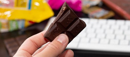 Eine Hand hält ein großes Stück dunkle Schokolade über einer Computertastatur, die von leeren Süßigkeitentüten umsäumt ist.