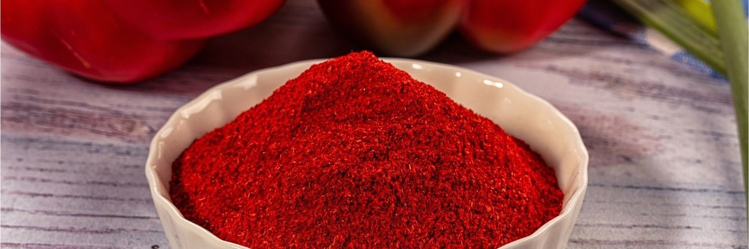 Auf einem Holztisch steht eine weiße Porzellanschüssel mit gemahlenem Paprikapulver. Im Hintergrund liegen zwei rote Paprikaschoten.