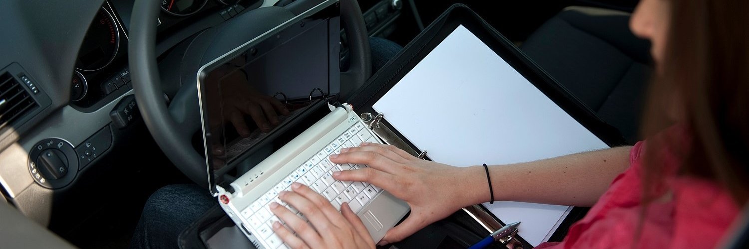 Eine Frau sitzt am Steuer eines Wagens, auf dem Schoß eine Schreibmappe und ein Laptop auf dem sie schreibt. 