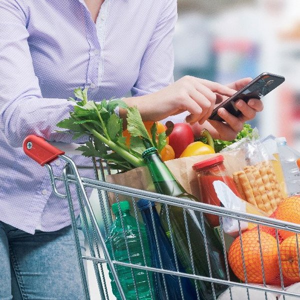 Eine Frau mit einem Handy in der Hand lehnt auf einem Einkaufswagen mit Lebensmitteln.