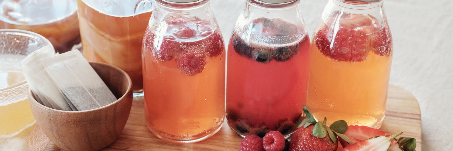 Auf einem Holzbrettchen stehen drei Schraubdeckelfläschchen mit einer sprudelnden klaren Flüssigkeit und Himbeeren, Blaubeeren und Erdbeeren. Daneben liegen einzelne Früchte und Teebeutel. 