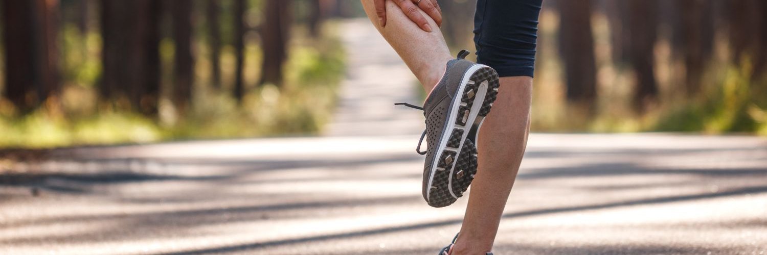 Eine Joggerin auf einem Waldweg hält ihre Wade mit der Hand. 