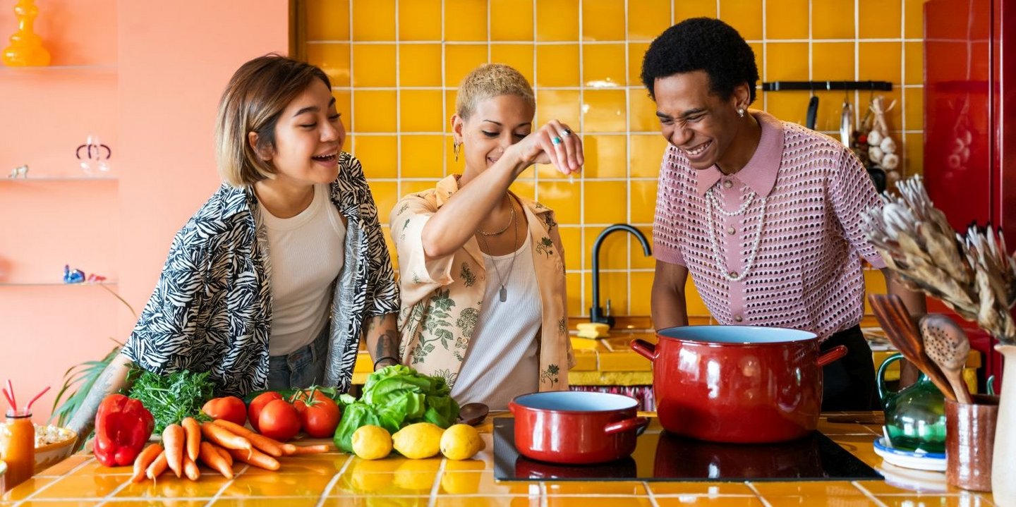 Drei Personen stehen am Herd, eine Person streut Salz in Kochtopf.