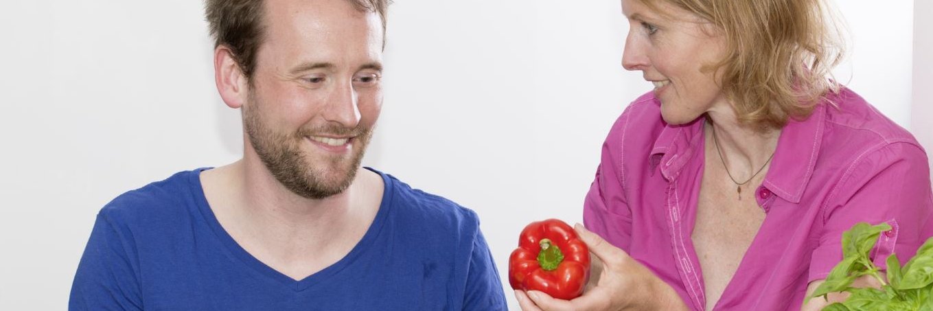 Eine Mann und ein Frau sitzen am Tisch mit Lebensmittel. Die Frau hält eine Paprika in ihrer Hand. Der Mann schneidet Gemüse.