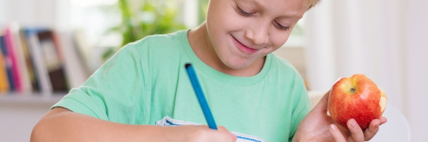 Ein blonder Junge im Grundschulalter sitzt an einem Tisch und schreibt in ein Heft. In der Hand hält er einen angebissenen Apfel.