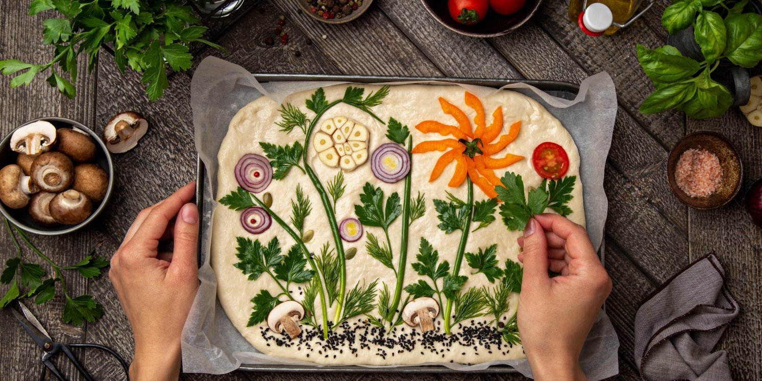 Auf einem dunklen Tisch steht ein Backblech mit Backpapier und ausgerolltem Focacciateig. Eine Frau dekoriert darauf Pflanzenmotive mit Zwiebeln, Kräutern, Knoblauch, Paprika, Pilzen und Tomaten.. Artikel "Herzhafte und süße Focaccia" öffnen.