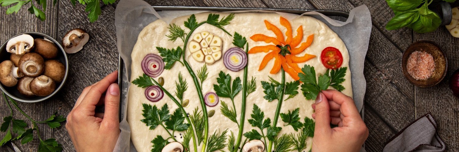 Auf einem dunklen Tisch steht ein Backblech mit Backpapier und ausgerolltem Focacciateig. Eine Frau dekoriert darauf Pflanzenmotive mit Zwiebeln, Kräutern, Knoblauch, Paprika, Pilzen und Tomaten.