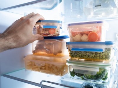Eine Hand greift in einen Kühlschrank, in dem Boxen mit gekochten Lebensmitteln gelagert sind.