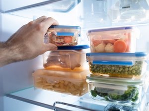Eine Hand greift in einen Kühlschrank, in dem Boxen mit gekochten Lebensmitteln gelagert sind.