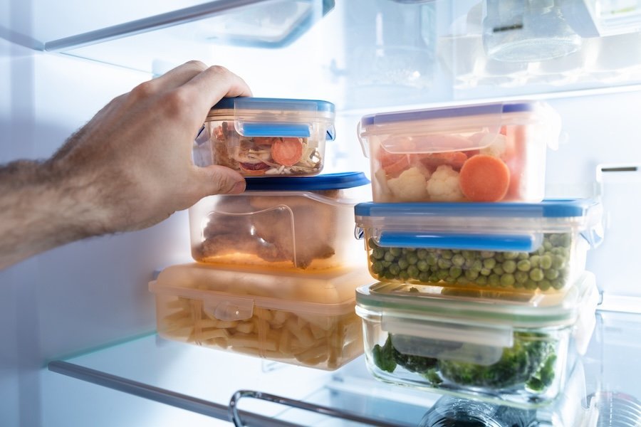 Eine Hand greift in einen Kühlschrank, in dem Boxen mit gekochten Lebensmitteln gelagert sind.