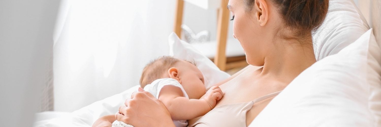 Eine junge Frau sitzt zuhause im Bett und stillt einen Säugling.