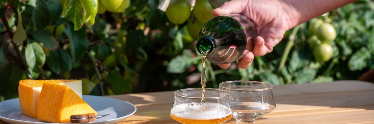 Auf einem Tisch im Freien unter einem Apfelbaum mit Früchten an den Zweigen stehen zwei Gläser und ein Teller mit Käse. Eine Hand gießt Schaumwein aus einer Flasche in eines der Gläser.