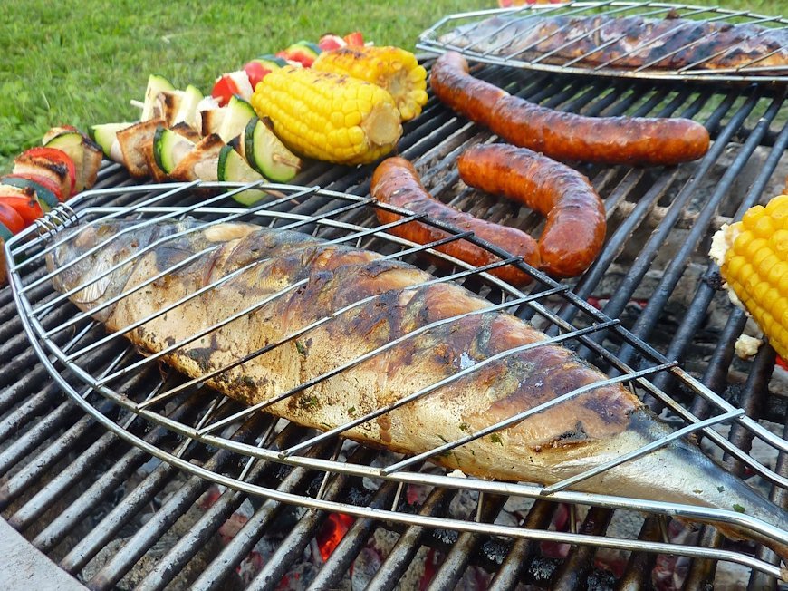 Ein ganzer Fisch in einer Grillzange liegt auf einem Grillrost mit Würstchen und Gemüse.