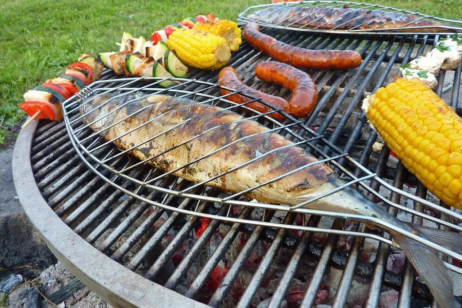 Ein ganzer Fisch in einer Grillzange liegt auf einem Grillrost mit Würstchen und Gemüse.