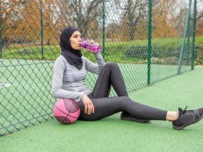 Eine Frau in Sportkleidung lehnt mit einem Ball am Zaun eines Spielfelds und trinkt aus einer Wasserflasche.