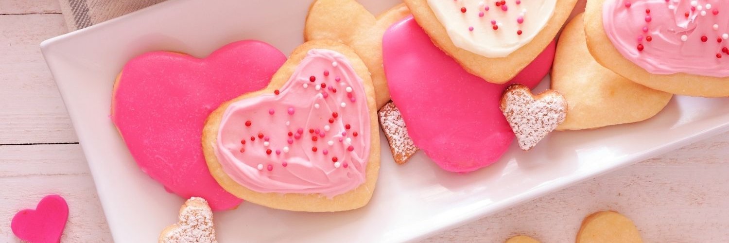 Auf einem Holztisch steht eine längliche weiße Schale mit herzförmigen Keksen mit pinker und rosafarbener Glasur und Zuckerperlen verziert.