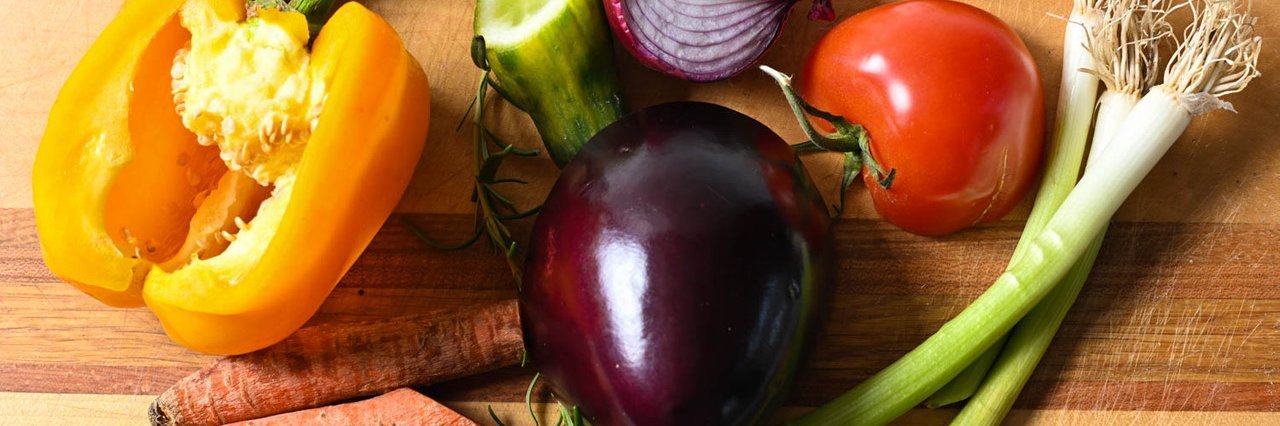 Verschiedene Gemüsesorten wie Paprika, Aubergine, Tomate, Möhre oder Zwiebel liegen teils angeschnitten nebeneinander auf einem Tisch.