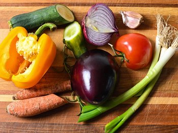 Verschiedene Gemüsesorten wie Paprika, Aubergine, Tomate, Möhre oder Zwiebel liegen teils angeschnitten nebeneinander auf einem Tisch.