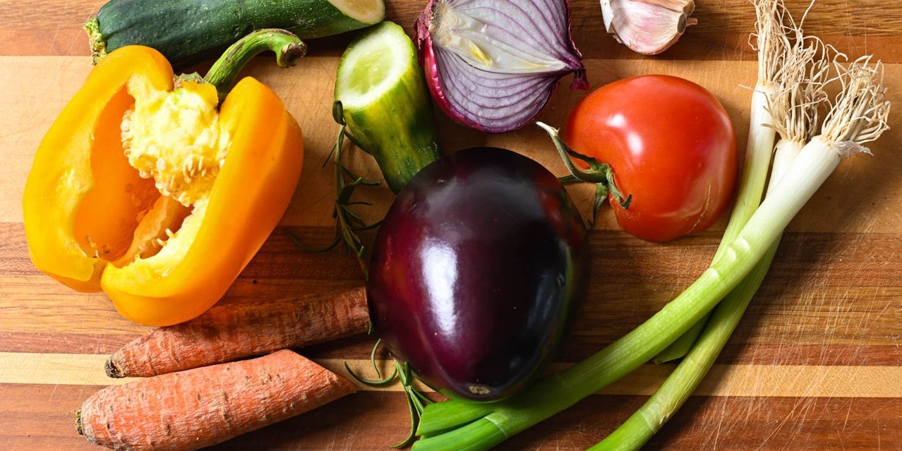 Verschiedene Gemüsesorten wie Paprika, Aubergine, Tomate, Möhre oder Zwiebel liegen teils angeschnitten nebeneinander auf einem Tisch.. Artikel "Wenn Essen im Müll landet" öffnen.