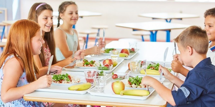 Zwei Jungen und drei Mädchen sitzen fröhlich an einem Tisch in der Mensa und essen gemeinsam ihr Mittagessen.. Artikel "Ernährungsbildung in den Ganztag bringen" öffnen.