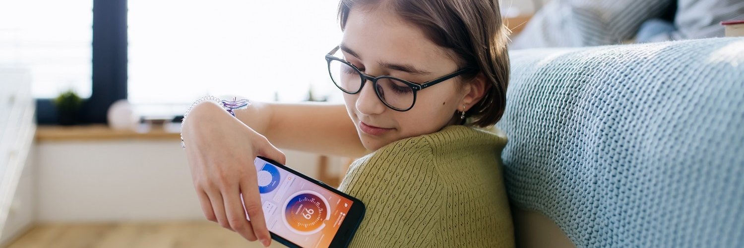 Eine junge Frau hat einen Blutzuckersensor am linken Oberarm. Sie hält ihr Handy an den Sensor, um mithilfe einer App ihren Blutzuckerspiegel checken zu können. 