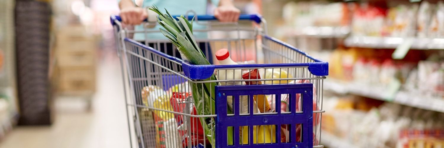 Eine Frau schiebt einen Einkaufswagen durch einen Supermarkt. Darin liegen Milch, Obst, Gemüse, Öl und Teigwaren.