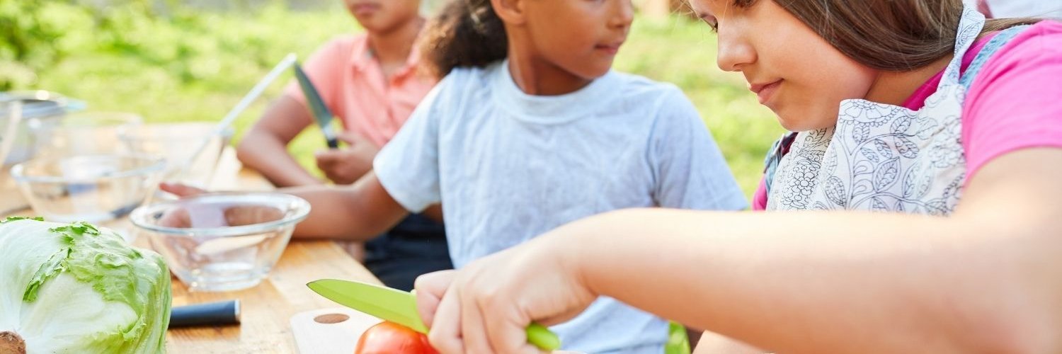 Mehrere Schulkinder sitzen an einem Tisch mit Lebensmitteln. Sie haben Schneidebretter, Schüsseln und Messer vor sich. Ein Mädchen mit Schürze schneidet eine Tomate.