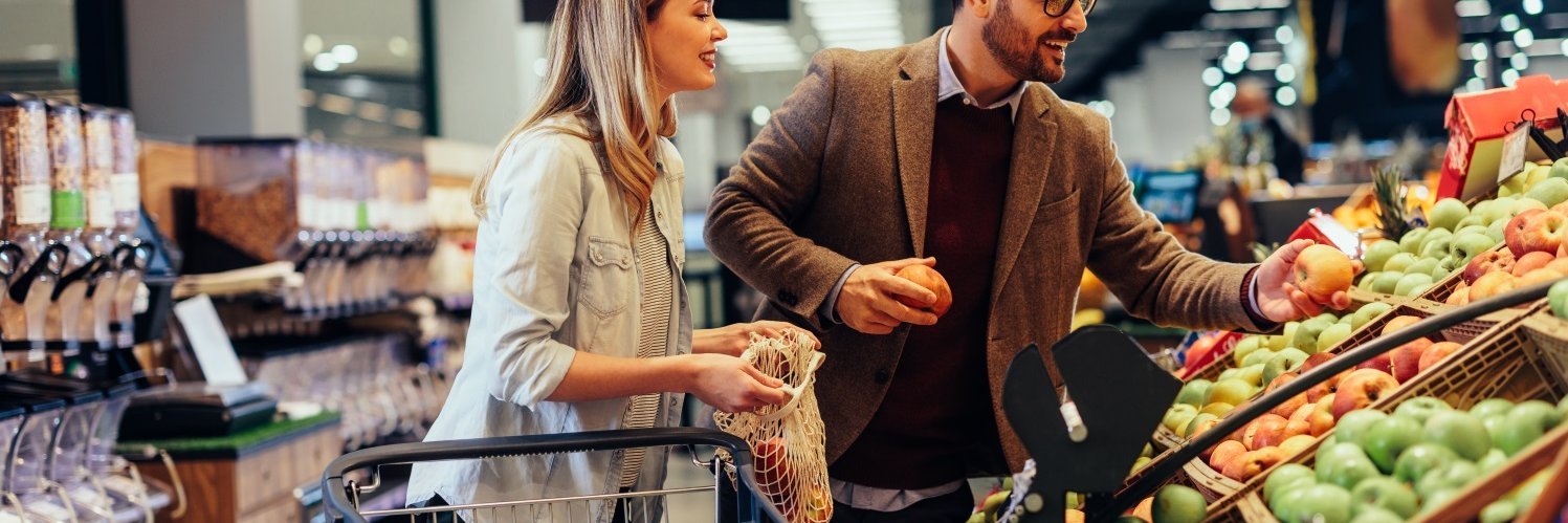 Ein Mann und eine Frau mittleren Alters stehen im Supermarkt an der Obst- und Gemüsetheke, neben sich einen Einkaufswagen. Der Mann hält Äpfel in den Händen und die Frau ein geöffnetes Gemüsenetz.