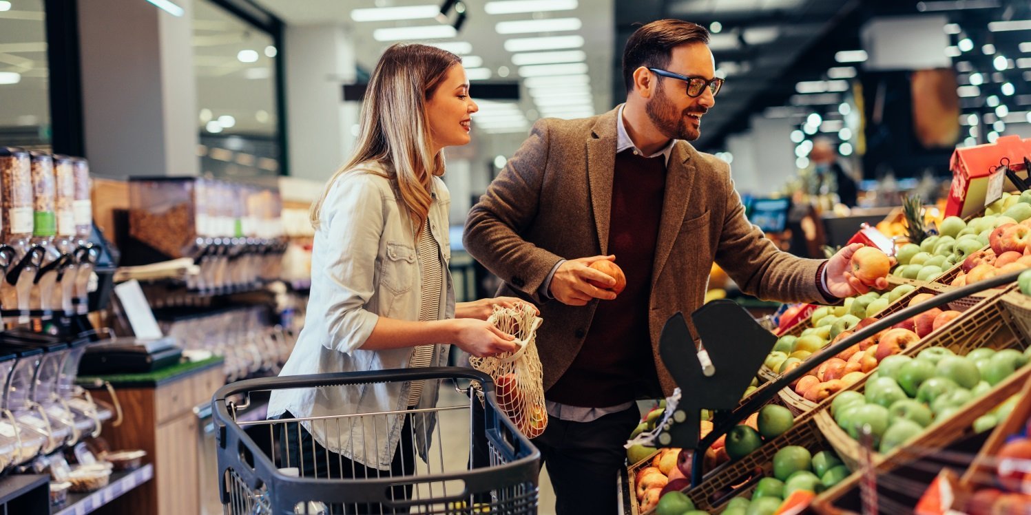 Ein Mann und eine Frau mittleren Alters stehen im Supermarkt an der Obst- und Gemüsetheke, neben sich einen Einkaufswagen. Der Mann hält Äpfel in den Händen und die Frau ein geöffnetes Gemüsenetz.. Artikel "Wie essen die Deutschen?" öffnen.