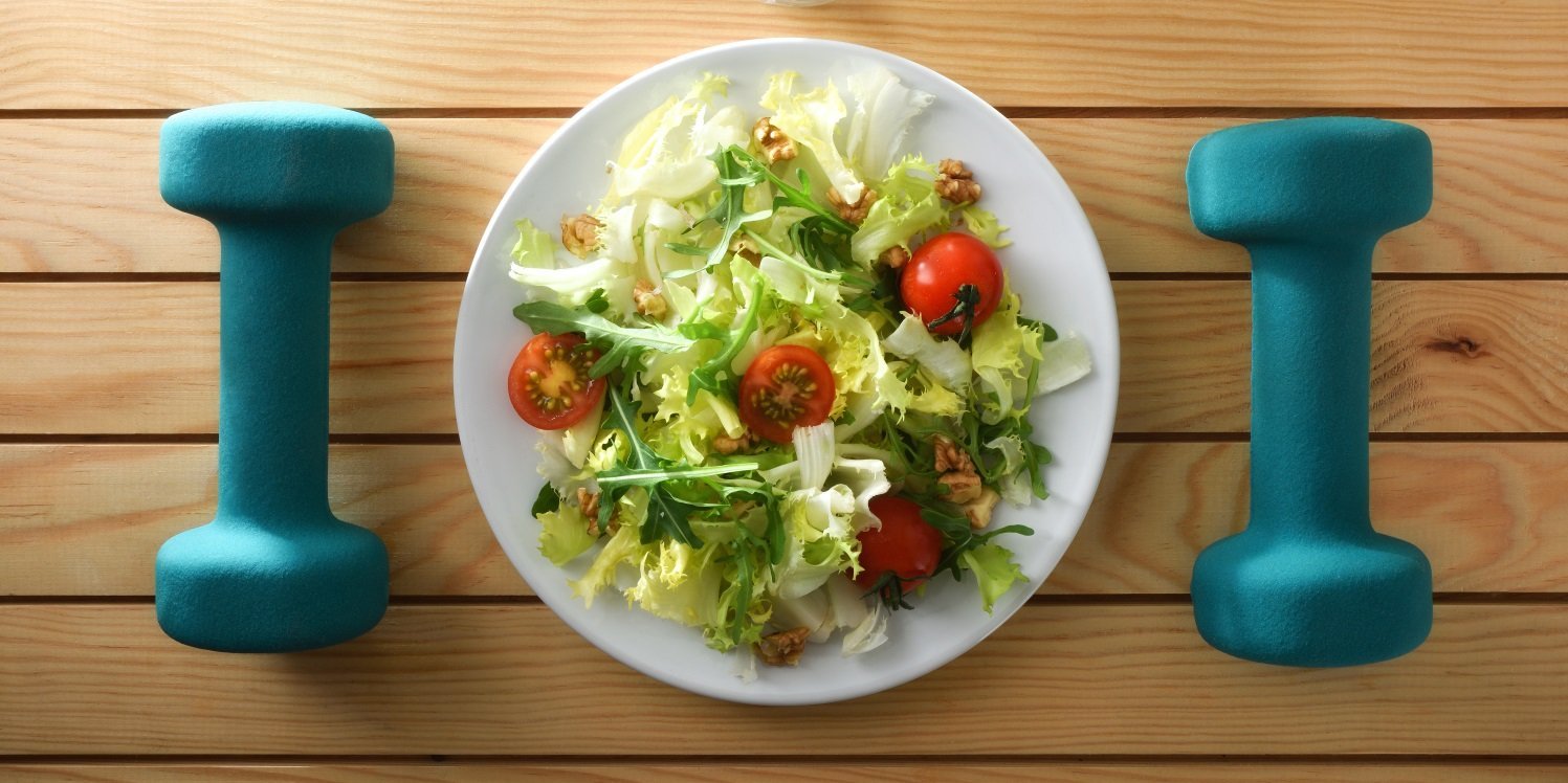 Zwischen zwei blauen Hanteln steht ein Teller mit Salat. . Artikel "Den Ursachen für Übergewicht auf der Spur" öffnen.