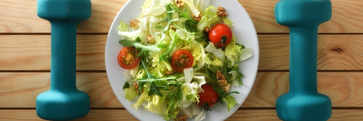Zwischen zwei blauen Hanteln steht ein Teller mit Salat. 