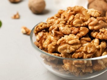 Auf einem hellen Untergrund steht eine Glasschale mit halbierten Walnusskernen. Im Hintergrund liegen Walnüsse mit Schale und einige Blätter.