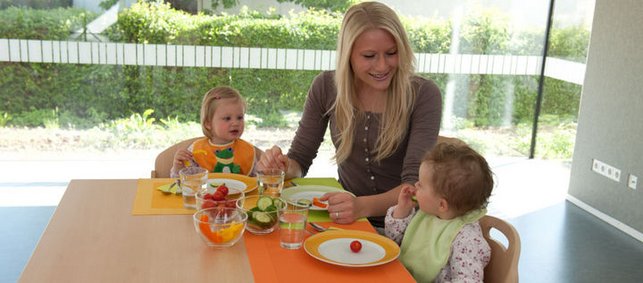 Zwei Kleinkinder sitzen unter Aufsicht einer Frau am Tisch und essen Gemüse