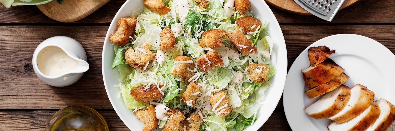 Auf einem dunklen Holztisch steht ein weißer Teller mit Blattsalat, Croutons und Käseraspeln, daneben ein Kännchen mit Dressing und Öl, ein Teller mit Hühnerbrust und ein Brett mit Käse und einer Reibe.