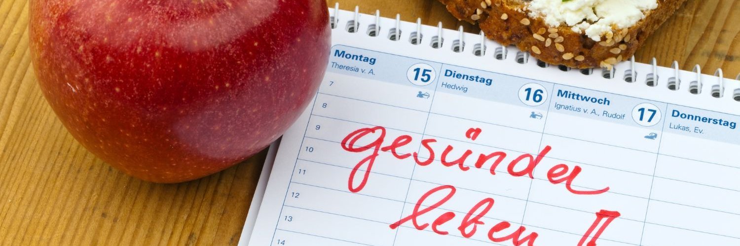 Auf einem Holztisch liegt ein Kalender mit dem Eintrag „Gesünder leben“, daneben ein roter Apfel und ein Vollkornbrot mit Frischkäse, Radieschen, Gurke und Schnittlauch belegt.